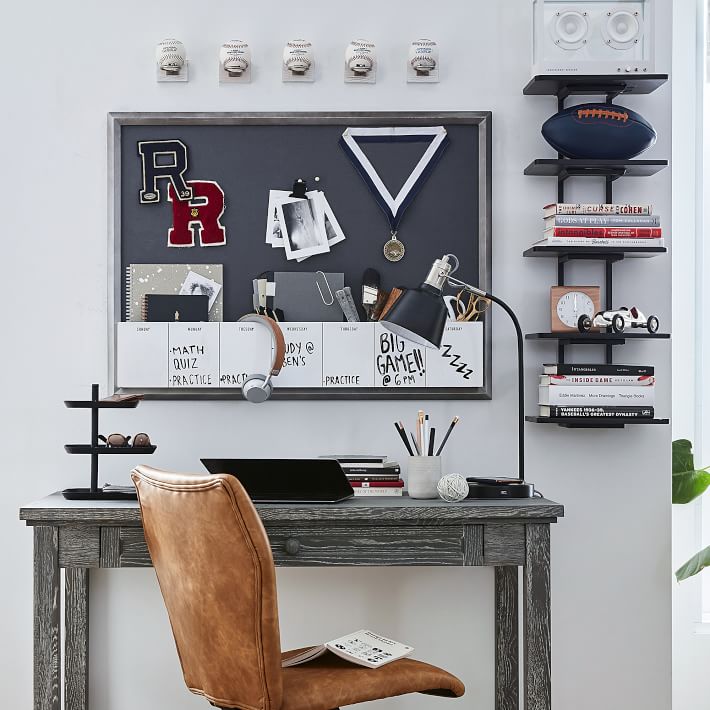 Desk with leather chair, lamp, and organised supplies. Noticeboard displays notes, medals, and sports items. Shelves hold books, rugby ball, and speakers against a white wall.
