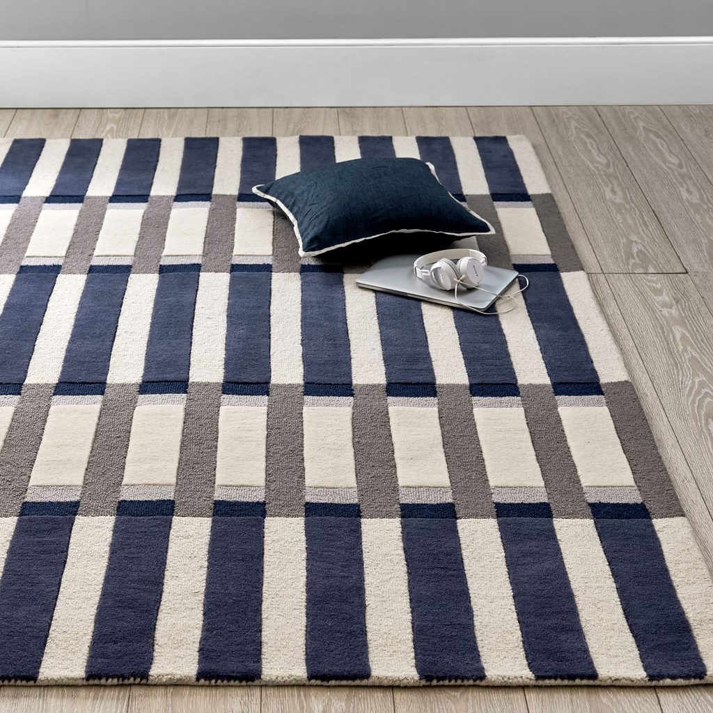 A striped rug with navy, grey, and cream colours lies on a wooden floor. A dark cushion, tablet, and white headphones are placed on the rug.
