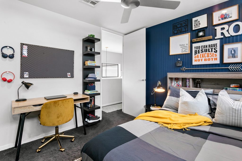 Modern bedroom with a bed, yellow chair at a desk, wall shelves, framed quotes, and a ceiling fan; bed has grey, black, and yellow bedding.