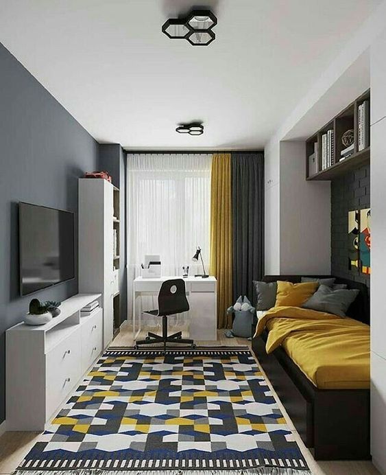 Modern bedroom with a patterned rug, bed with yellow bedding, wall-mounted TV, white desk by a window, and shelves above the bed.