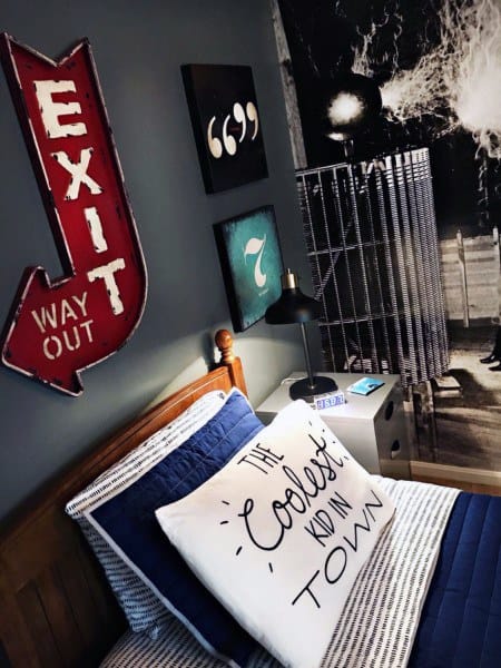 A bed with a pillow that says “The Coolest Kid in Town,” a red EXIT sign, wall art, and a desk lamp in a modern, styled bedroom.
