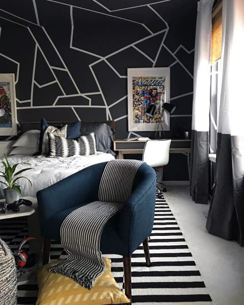 Contemporary bedroom with geometric black walls, striped rug, blue chair with a striped throw, bed with patterned cushions, and a desk with framed comic art.