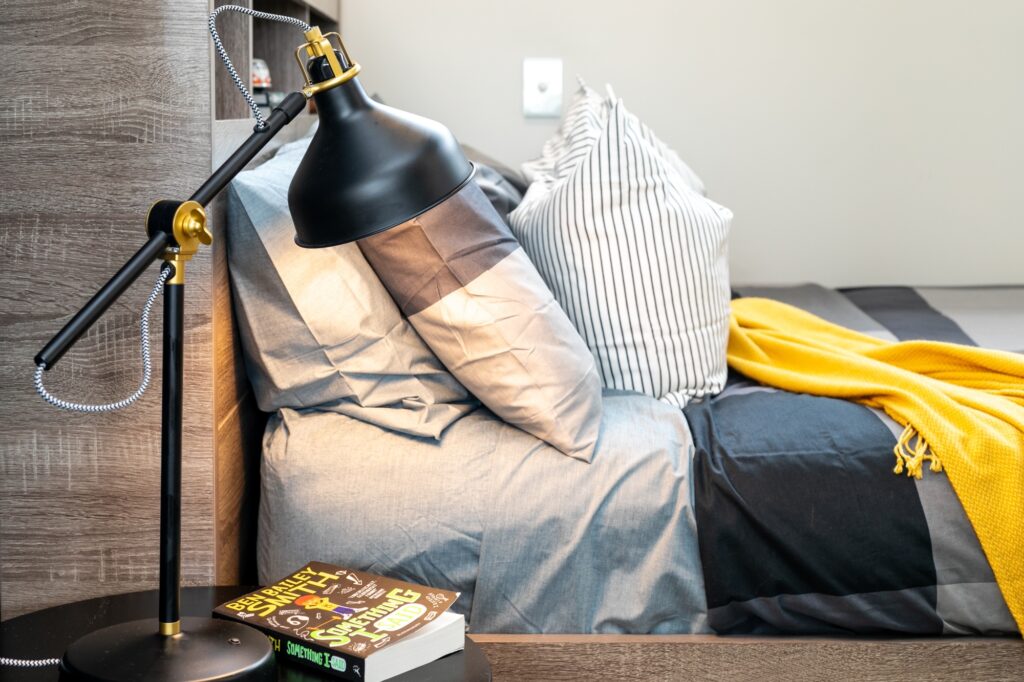 A modern bedside with a black desk lamp, two books, and a neatly made bed with grey sheets, striped pillows, and a yellow blanket.