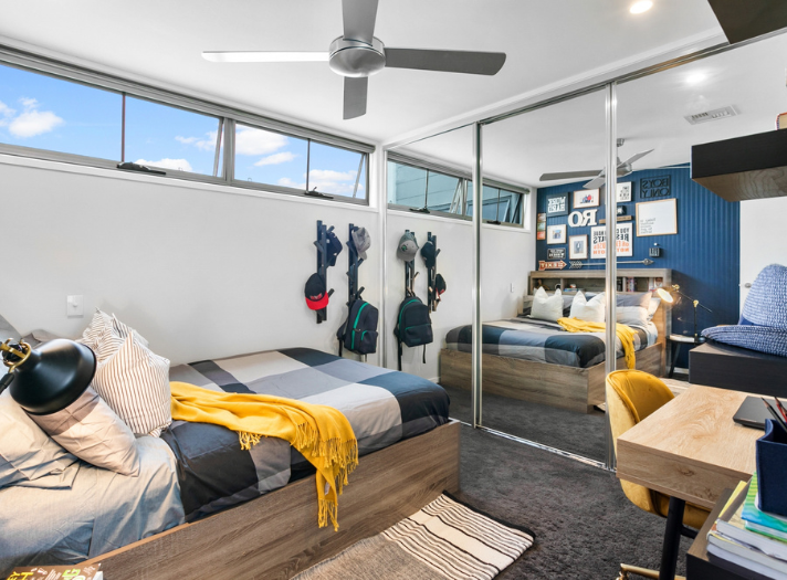 Modern bedroom with a single bed, yellow blanket, mirrored wardrobe doors, desk, chair, wall shelves, hats on hooks, and a ceiling fan.