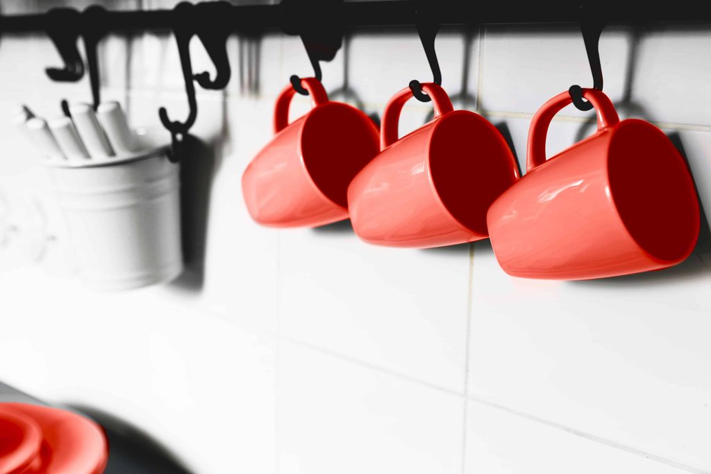 Three red ceramic mugs hang from black hooks on a white tiled kitchen wall, with a white container holding utensils visible in the background.