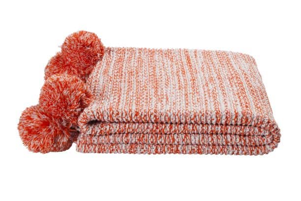 Folded orange and white knitted blanket with three large pompoms on one end, isolated on a white background.