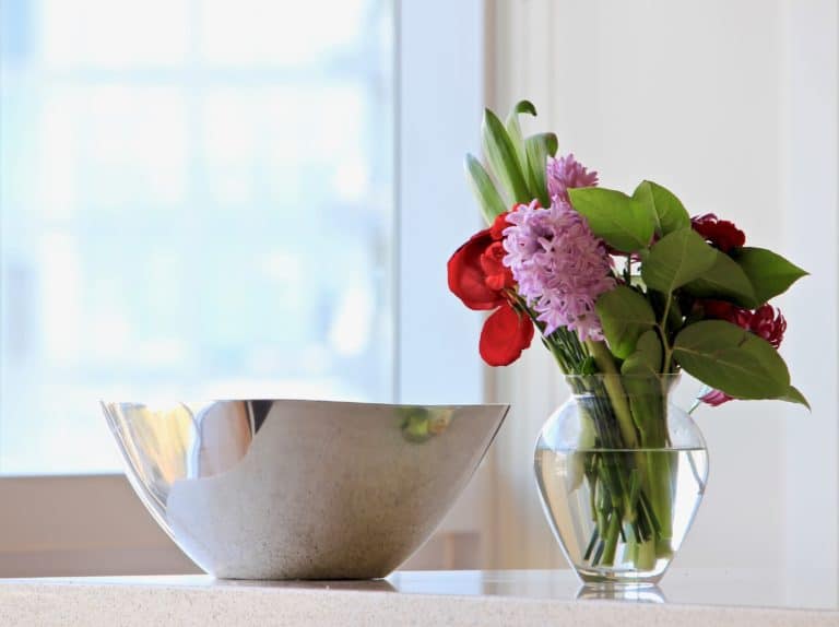 A metal bowl and a glass vase with red and purple flowers and green leaves sit on a white surface near a bright window.