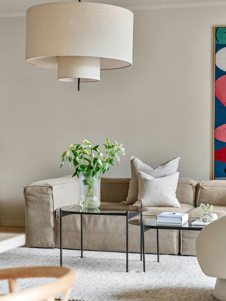 A beige corner sofa with cushions sits behind a glass vase of green foliage on a black coffee table, under a large layered beige pendant light. A colourful abstract painting hangs on the wall.