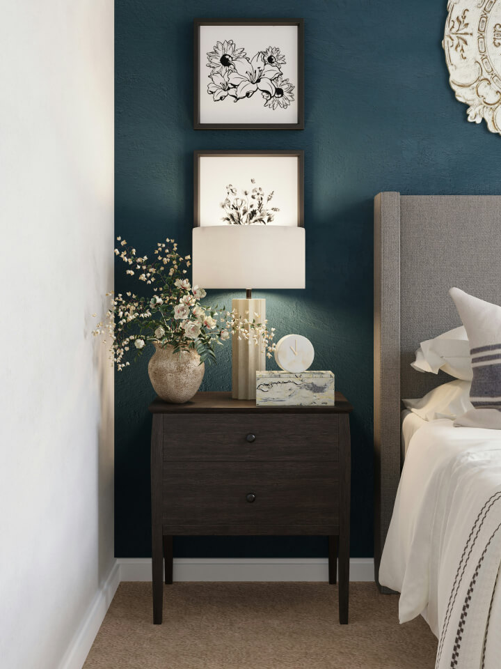 A dark wood bedside table with a lamp, flower vase, and decorative items beside a bed with a grey headboard, set against a teal wall with framed artwork.