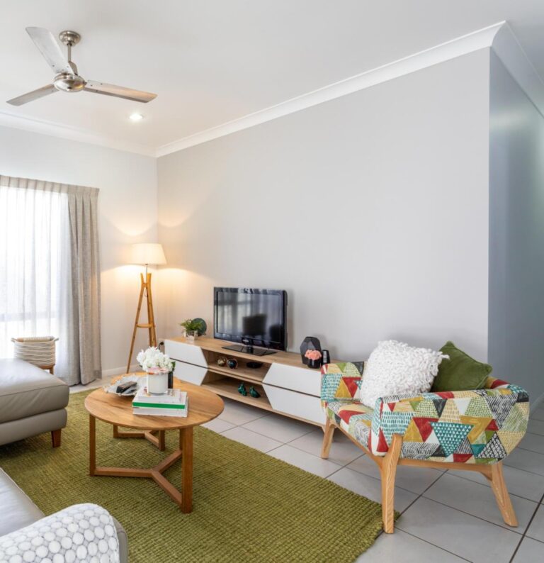 Modern living room with a patterned armchair, round wooden coffee table, TV on a low console, a floor lamp, grey sofa, green rug, and tiled floor.