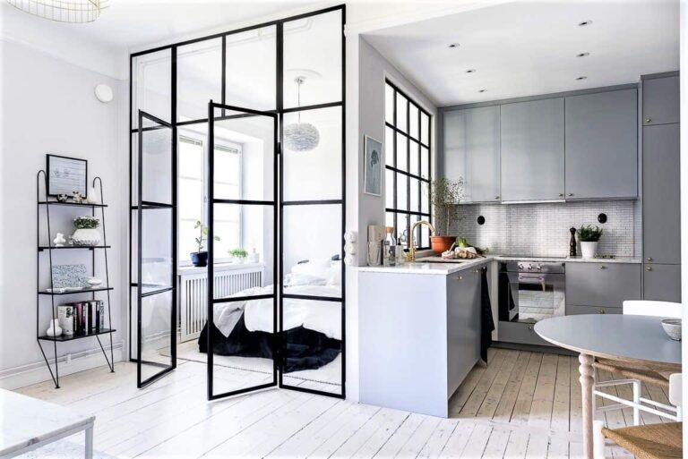 Modern studio flat with light wood floors, glass partition separating the bedroom, small kitchen with grey cupboards, and minimalistic decor.