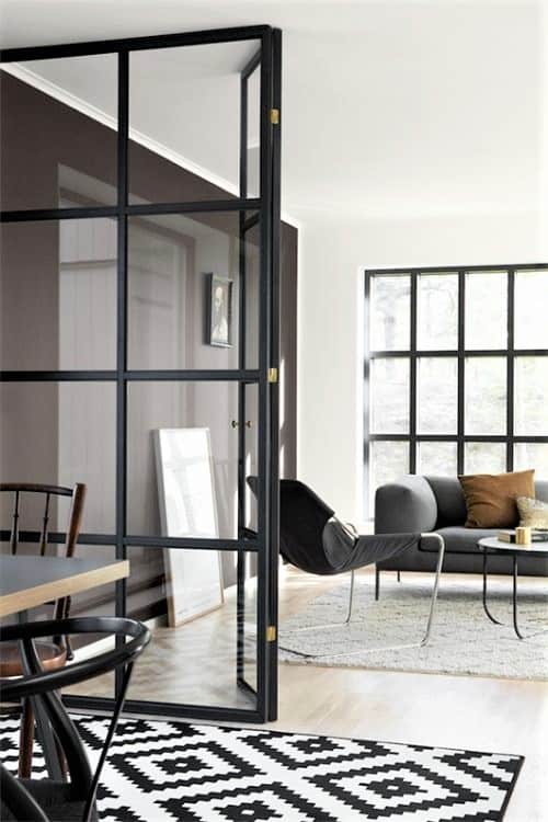 Modern living room with black-framed glass door, grey sofa, black chair, round coffee table, and geometric black-and-white rug.