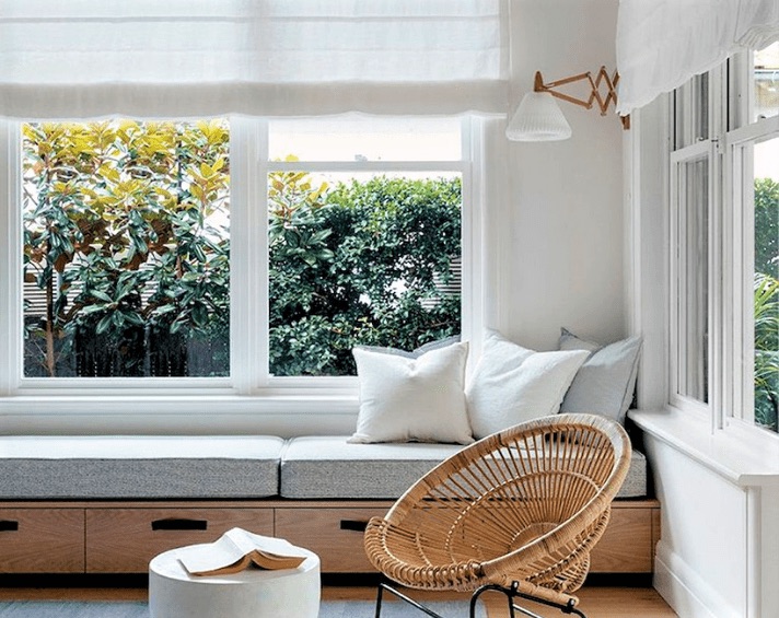 A bright room with large windows, a built-in bench with cushions, a wicker chair, a small white table, and greenery visible outside.