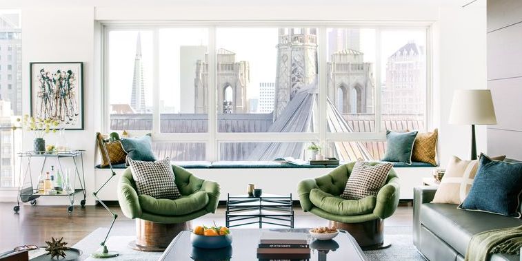 Modern living room with two green armchairs, a window seat, cityscape view, coffee table, and decorative accents. Large windows let in natural light.