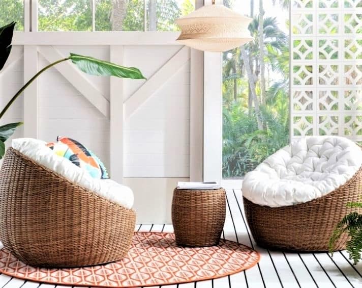 Two cushioned rattan chairs and a matching side table are arranged on a round rug in a bright, tropical indoor space with large windows and greenery outside.