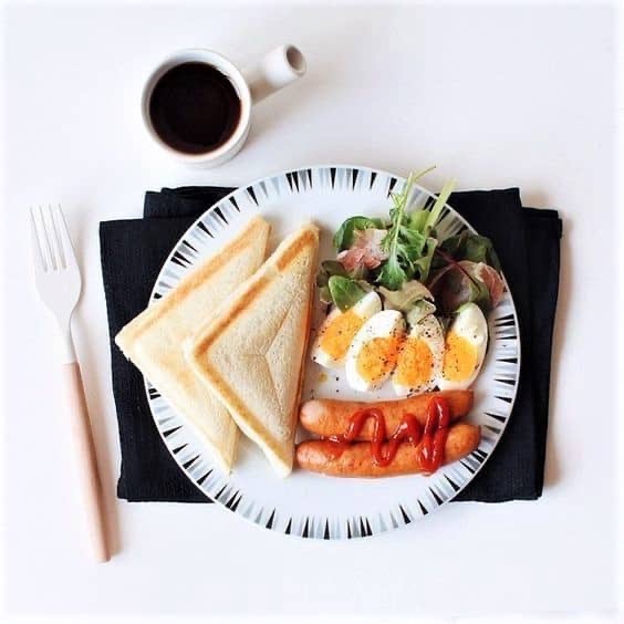 Plate with toast, salad, sliced boiled eggs, sausages with tomato sauce, and a cup of coffee on a white table with a fork and black serviette.