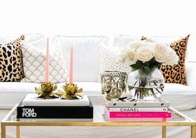 A glass coffee table holds books, candles, a gold Buddha head, and a vase of white roses. White sofa with patterned cushions is in the background.