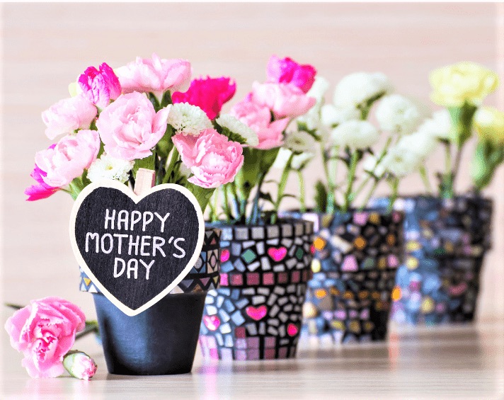 Four flower pots filled with pink and white carnations, one with a heart-shaped “Happy Mother’s Day” sign in front, arranged on a light surface.