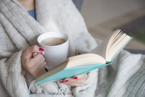 Person wrapped in a blanket holds a white mug of coffee or tea in one hand and an open book in the other.