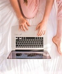 Person sitting cross-legged on a bed, using a laptop placed in front of them, with hands on the keyboard.