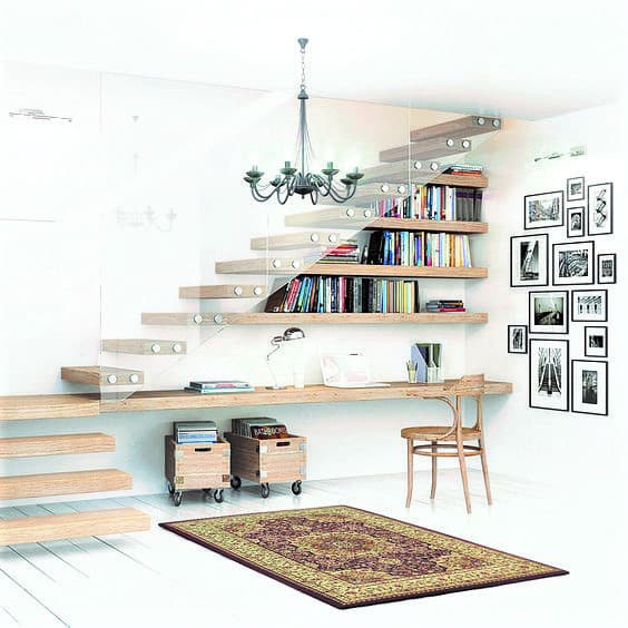 A modern study area under floating stairs with bookshelves, a wooden desk and chair, wall art, and a red patterned rug. Two storage boxes are placed under the desk.