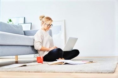 Woman sitting on the floor by a sofa, working on a laptop with notebooks, papers, and a red mug on a rug in a bright living room.