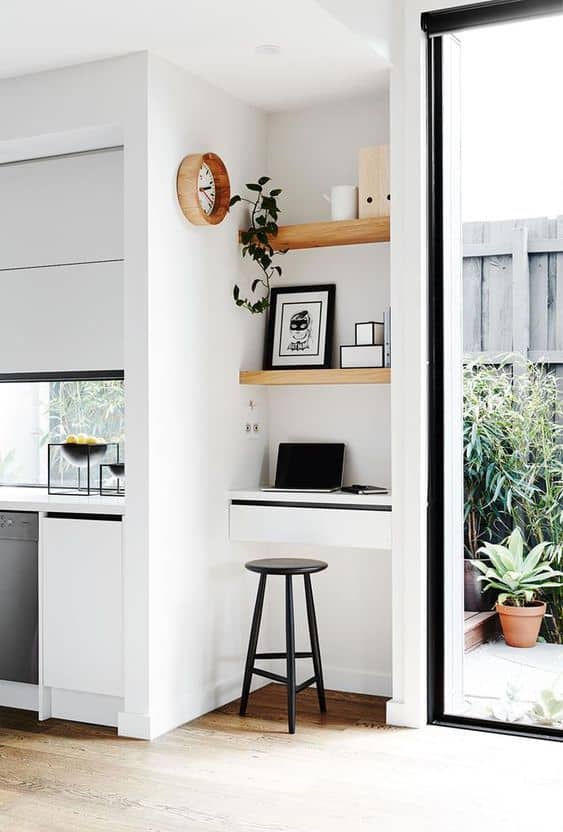 Minimalist home office nook with a wall-mounted desk, black stool, shelves with decor, and a laptop, next to a large glass door and indoor plants.