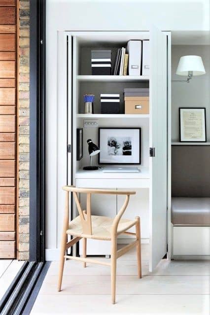 A small home office setup inside a white cupboard features shelves with boxes and books, a framed photo, a desk lamp, pens, and a wooden chair.