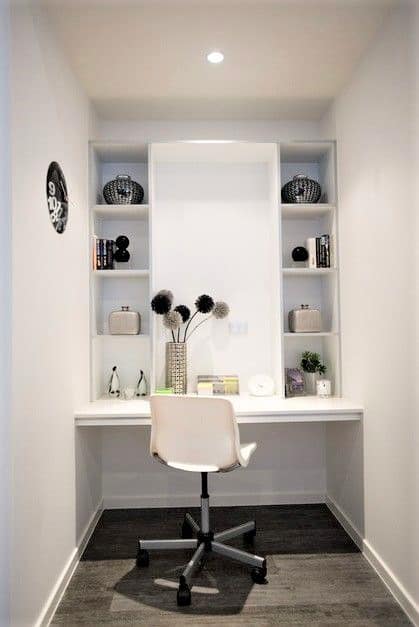 Small home office nook with a white desk, white chair, open shelving, decorative items, books, and a wall clock on the left; dark floor, white walls, and overhead lighting.