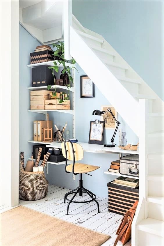 Small home office setup under white stairs with shelves, boxes, art supplies, a plant, a wooden swivel chair, a desk lamp, and framed pictures against a blue wall on white wooden floor.