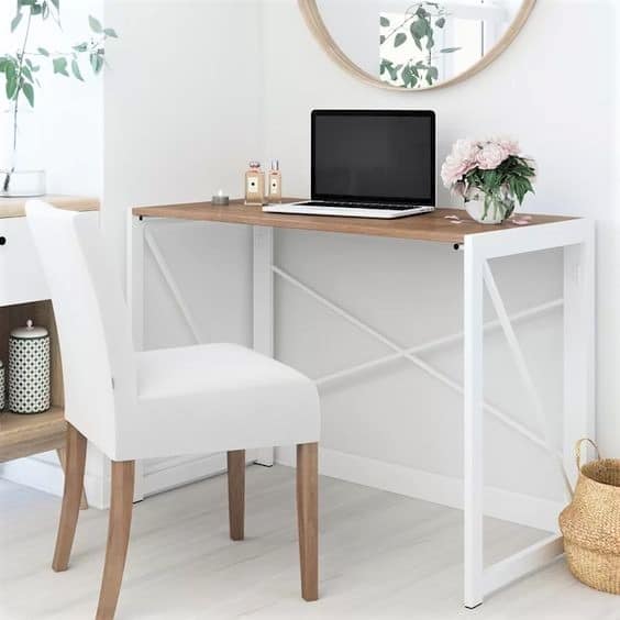 A small modern desk with a laptop, flowers, and perfume bottles sits in a bright room with a white chair and a round wall mirror above.