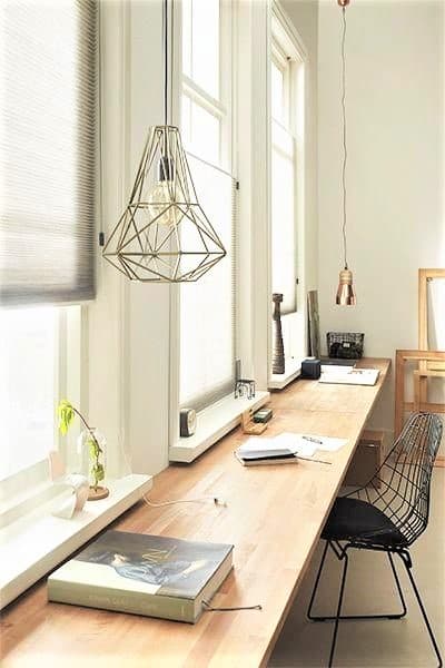 Minimalist workspace with a long wooden desk, wire chair, pendant lights, books, and décor by tall windows.