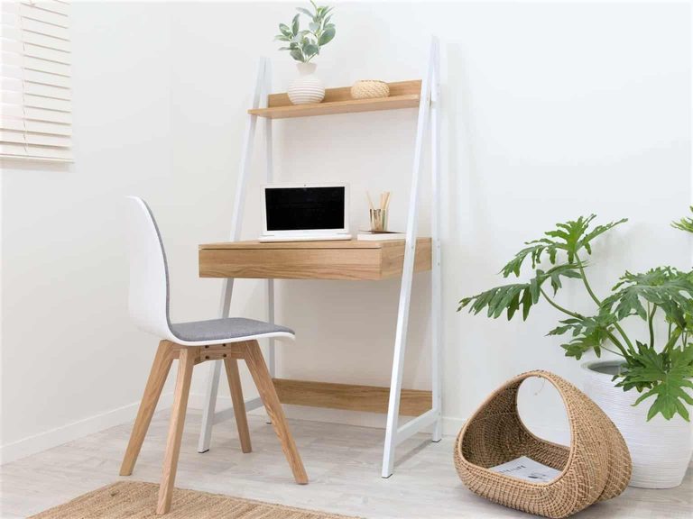 Small workspace with a wooden desk, white chair, laptop, potted plants, and a woven basket on the floor in a minimalist room.