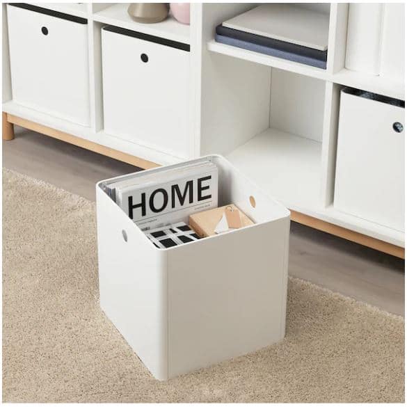 A white storage box on a beige carpet holds books and miscellaneous items in front of a white cubby shelving unit.
