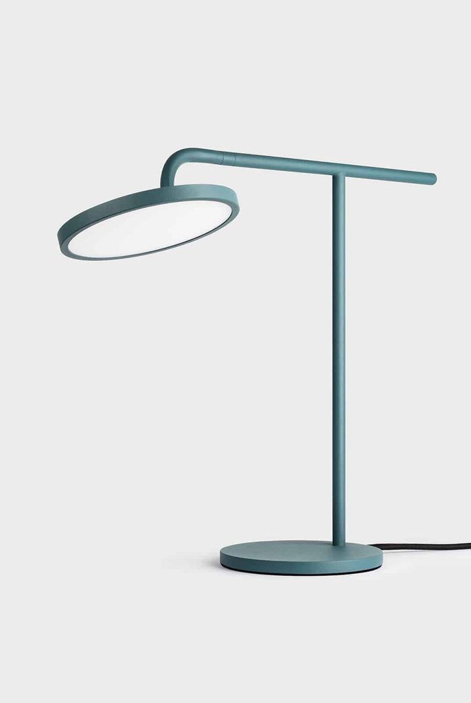 Modern teal desk lamp with a round, flat LED light, horizontal arm, vertical stand, and circular base, set against a plain light background. A black power lead extends from the base.