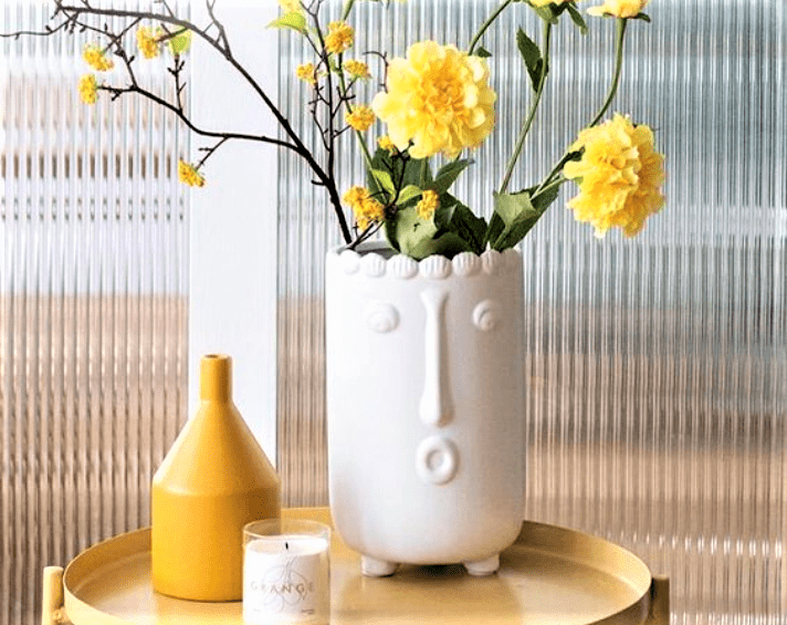 White vase with a face design holds yellow flowers, placed on a round yellow table with a yellow bottle and a candle nearby.