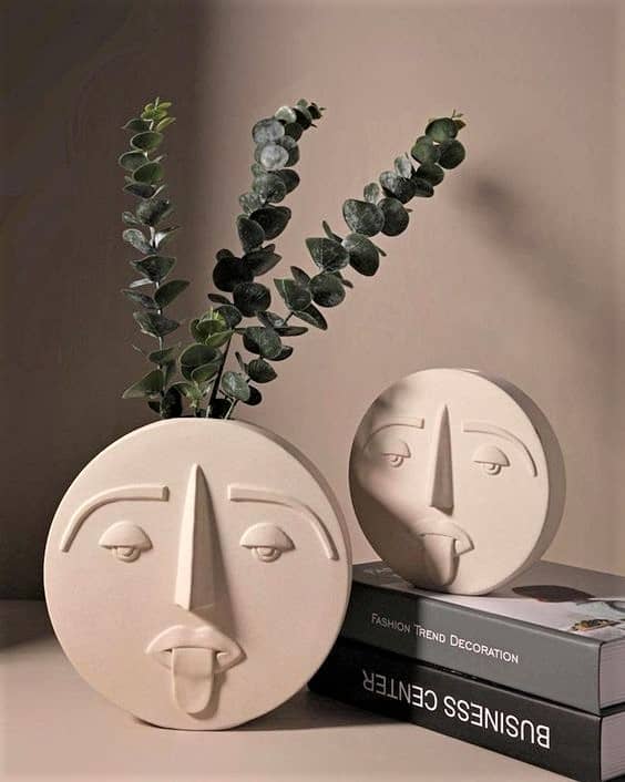 Round ceramic vase with a minimalist face design holding eucalyptus stems, next to a matching round mirror, both placed on stacked books against a beige wall.