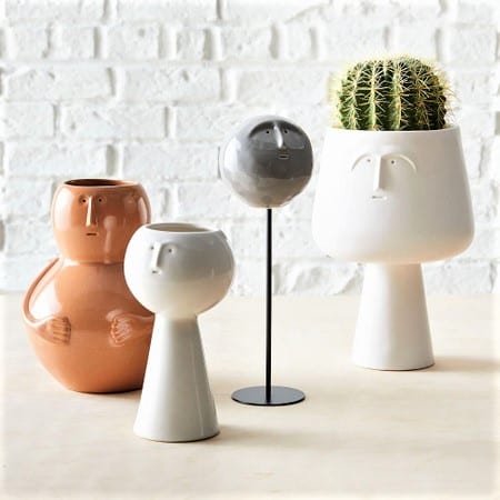 Four ceramic vases with minimal face designs; one white vase holds a cactus. The background is a white brick wall.