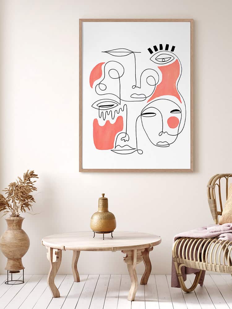 Framed abstract line art drawing with red accents hangs above a light wood table and wicker chair in a neutral-toned room.