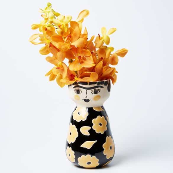 A ceramic vase shaped like a stylised human figure with a floral pattern holds a bouquet of orange-yellow flowers.