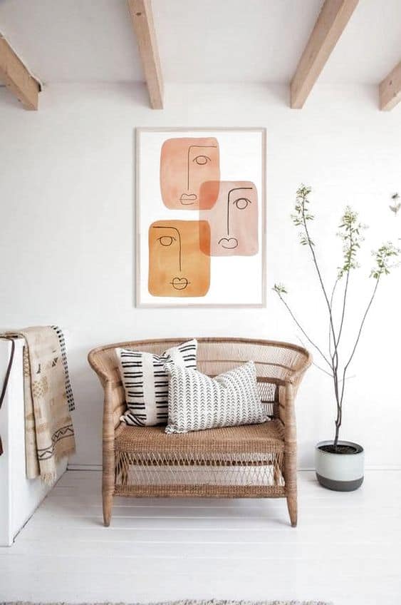 A wicker two-seater with patterned cushions sits beneath abstract face art on a white wall; a tall branch in a vase stands nearby in a bright, minimalist room with exposed wooden ceiling beams.
