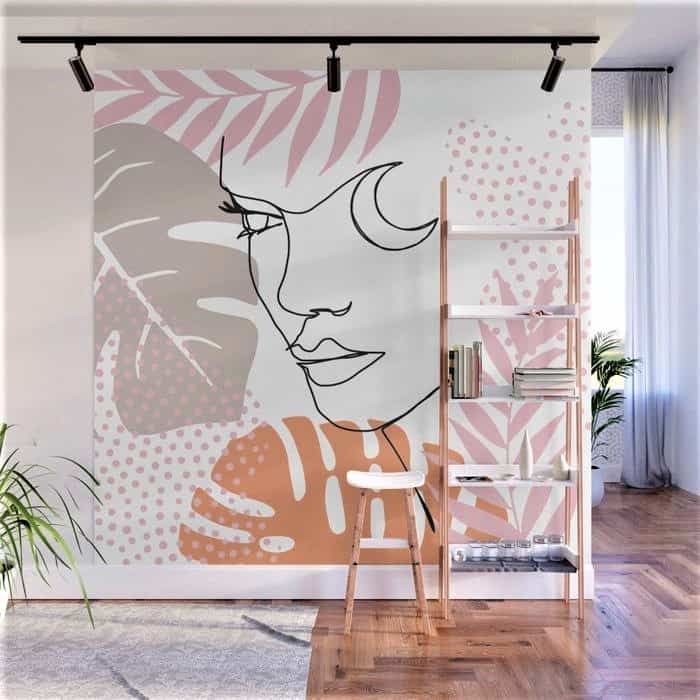 Modern living room with a large abstract mural of a face, botanical shapes, and dots in pink and beige tones behind a wooden bookcase and stool.