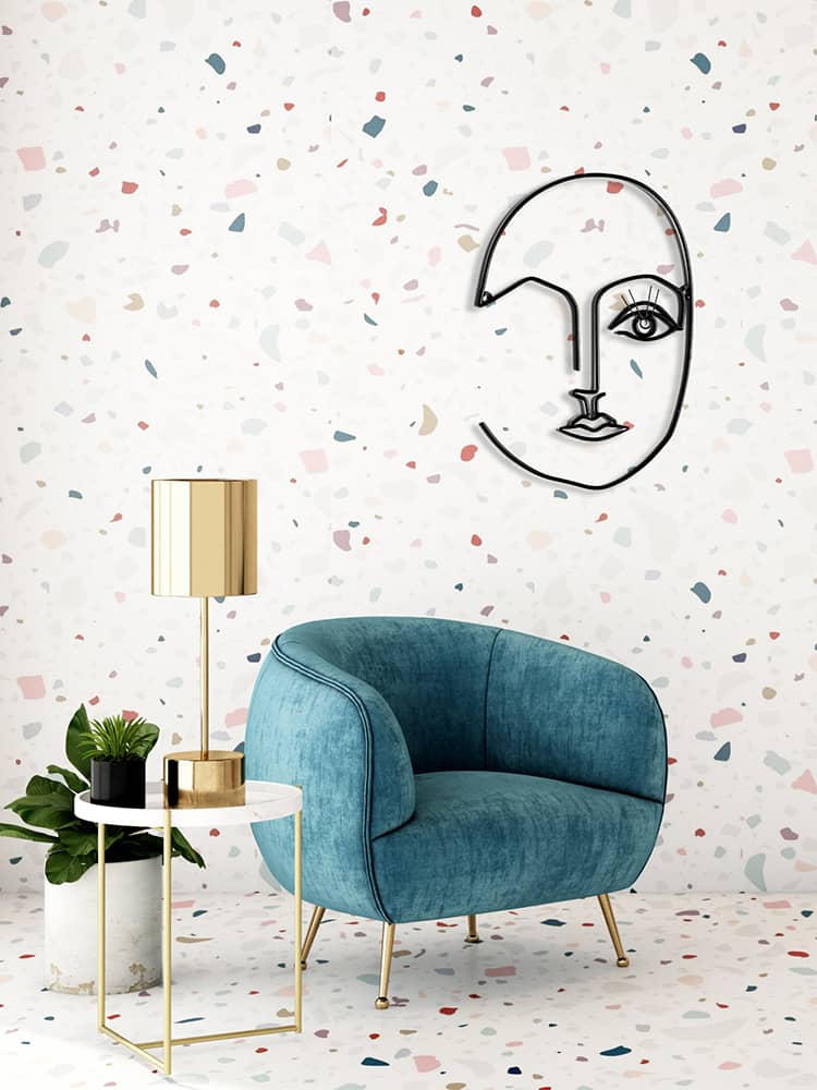 A blue velvet chair sits beside a gold lamp and small table on a terrazzo floor, with minimalist face wall art on a terrazzo-patterned wall.