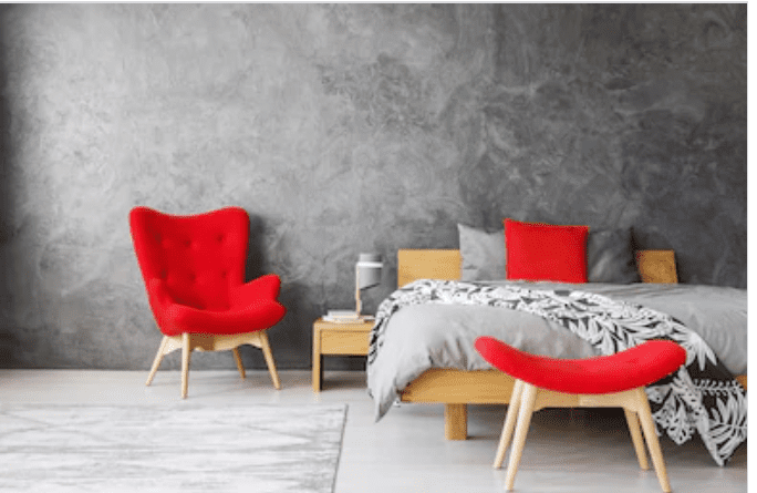 Modern bedroom with a grey textured wall, wooden bed and bedside table, grey bedding, and red accent chair, cushion, and bench.