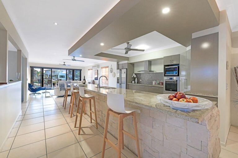 Modern kitchen with a large stone island, bar stools, stainless steel appliances, tiled flooring, and open plan leading to a bright living area. A bowl of fruit sits on the worktop.