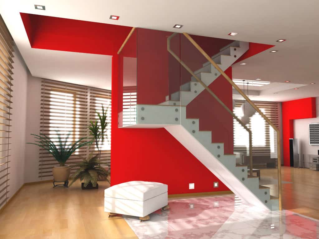 Modern interior with wooden floors, a glass and steel staircase against a red wall, a white ottoman, a potted plant, and large windows with horizontal blinds.