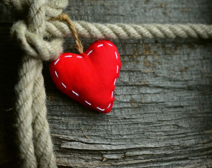 A red fabric heart with white stitching hangs from a thick rope tied in a knot against a weathered wooden background.
