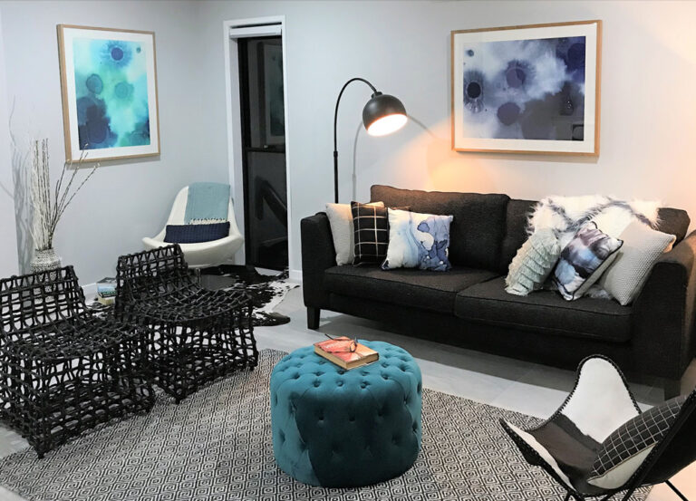 Modern living room with a dark sofa, blue tufted ottoman, black woven chairs, abstract art on the walls, a floor lamp, and neutral decor accents.