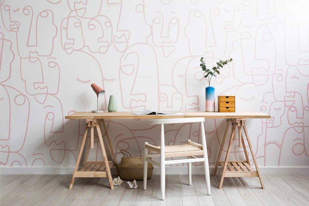 Wooden desk with a white chair, lamp, books, vases, and drawers against a white wall with abstract face line art.