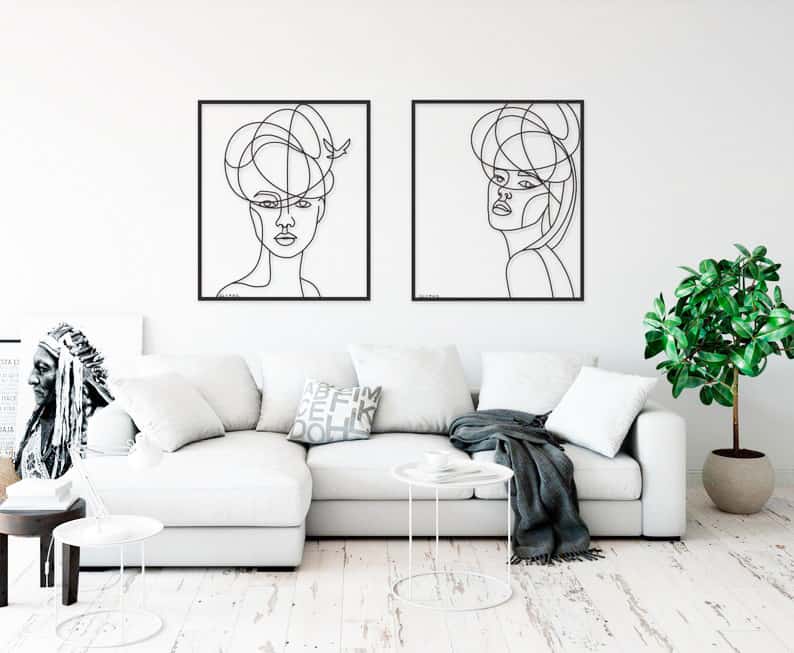 Modern living room with white corner sofa, two abstract line art portraits on the wall, a leafy plant, side tables, and a monochrome Native American headdress artwork on the left.